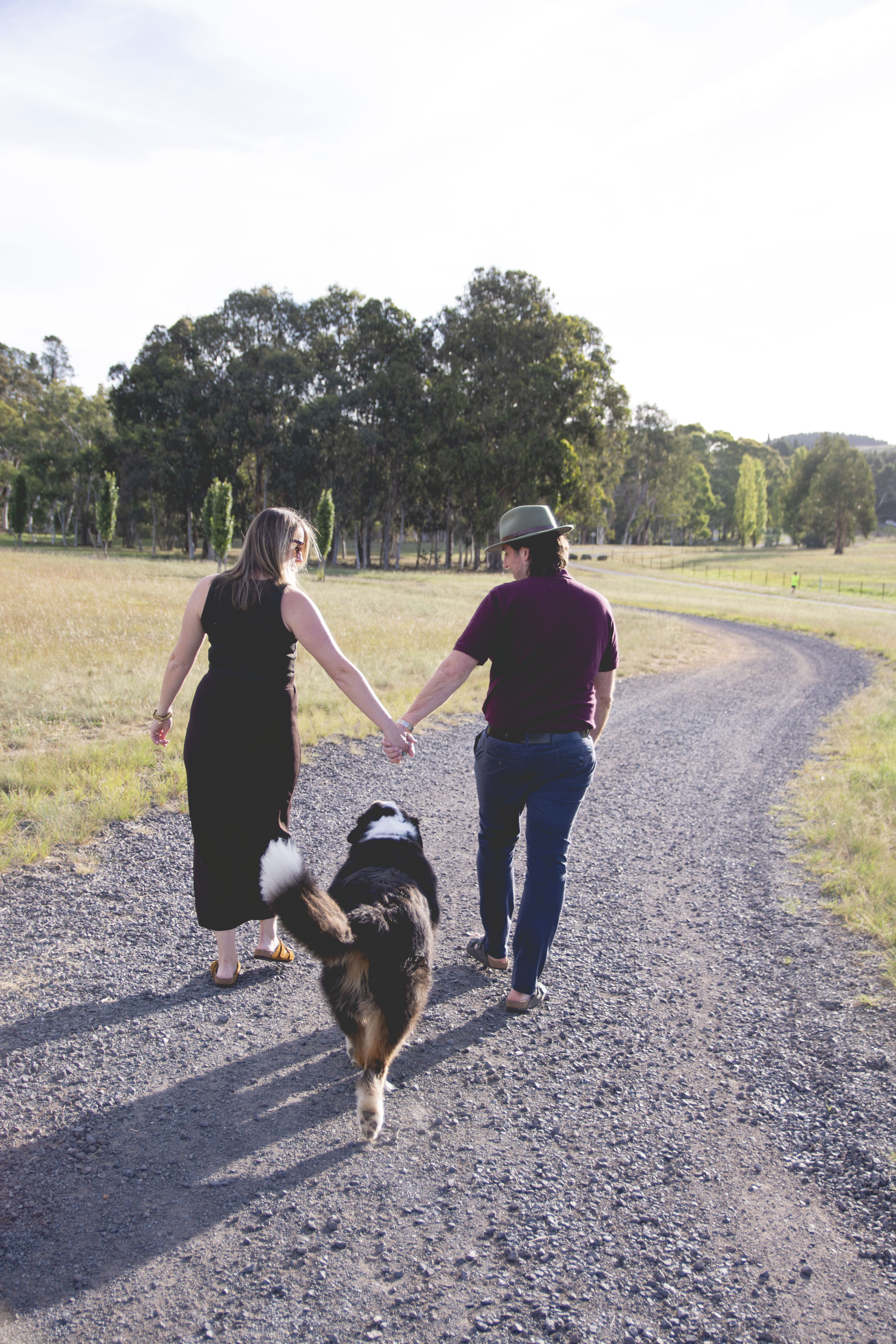 Image of a couple holding hands and walking on a winding gravel path, looking at their dog who walks between them